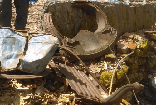 Rusty metal debris and old machinery parts scattered among leaves and stones in a wooded area.