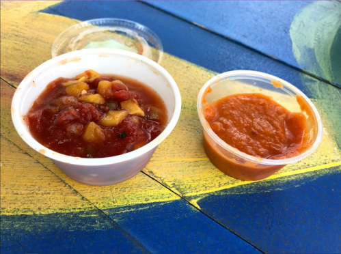Two small cups of salsa: one chunky with vegetables and the other smooth and orange, placed on a colorful table.