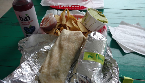 A wrapped burrito, chips with salsa, guacamole, and a drink on a green table. A note reads "No Dairy."
