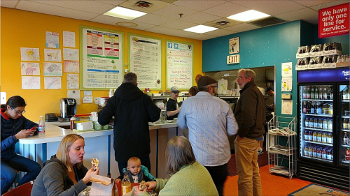 A busy café with customers at the counter, people seated, and colorful walls displaying menus and notices.