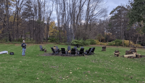A fire pit surrounded by black chairs in a grassy area, with a person standing nearby and trees in the background.