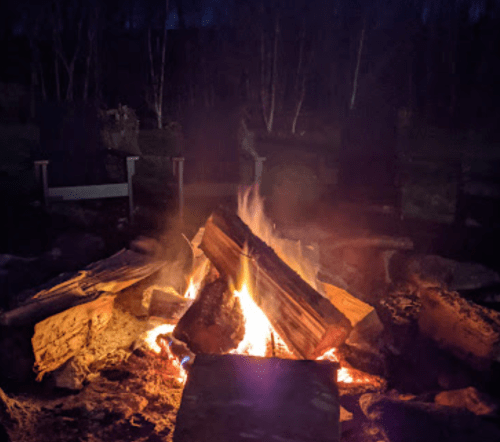 A cozy campfire burns brightly at night, surrounded by stone and wooden seating in a wooded area.