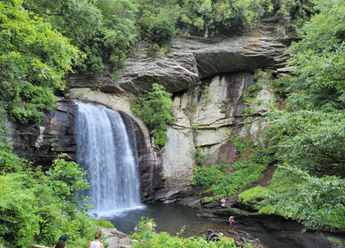 A serene waterfall cascades into a pool, surrounded by lush greenery and rocky cliffs, with people enjoying the scenery.