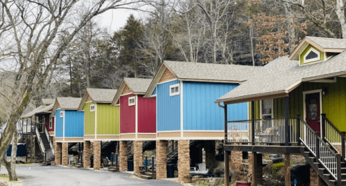 Colorful cabins lined along a tree-lined road, each elevated on stone pillars with a mix of blue, green, and red exteriors.