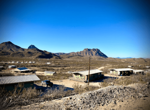 A desert landscape with small green houses, mountains in the background, and a clear blue sky.