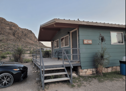 A blue house with a porch and steps, surrounded by mountains and desert vegetation, with a car parked nearby.