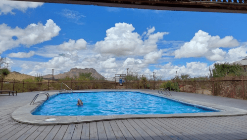 A serene pool surrounded by desert vegetation and mountains under a bright blue sky with fluffy white clouds.