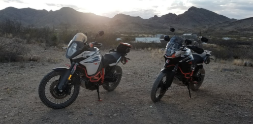 Two adventure motorcycles parked on a rocky terrain with mountains and a cloudy sky in the background.