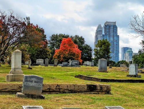 A cemetery with gravestones, colorful autumn trees, and a city skyline in the background under a cloudy sky.