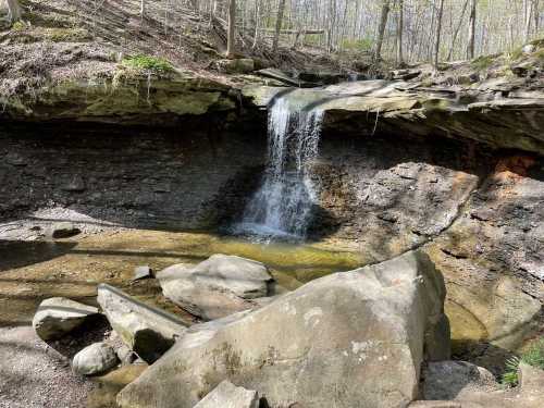 A serene waterfall cascades over rocks into a calm pool, surrounded by trees and natural stone formations.