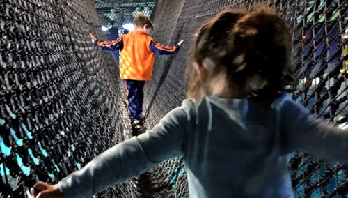 Two children play on a net bridge, one walking ahead in an orange shirt, while the other reaches out behind.