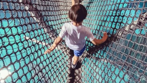A child walks carefully on a suspended net above a pool, with water visible below.