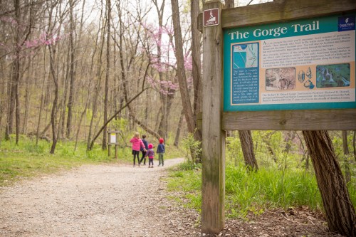 A family walks along a gravel path in a wooded area, with a sign for "The Gorge Trail" nearby.
