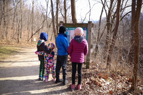 Four children in winter clothing stand by a trail sign in a wooded area, looking at the information displayed.