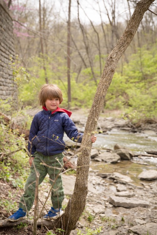 A young child stands by a creek, smiling while holding onto a tree, surrounded by greenery and rocky terrain.
