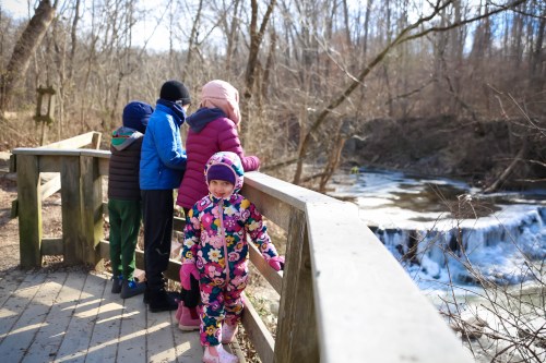 A group of children stands on a wooden platform by a river, with one girl in a colorful outfit smiling at the camera.