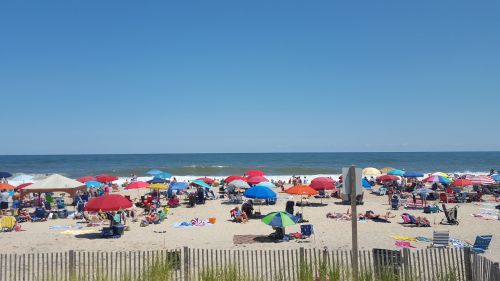 Crowded beach scene with colorful umbrellas, sunbathers, and waves under a clear blue sky.