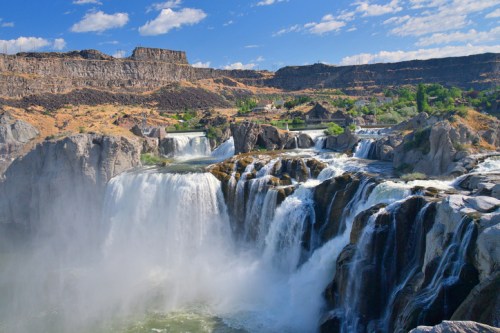 A stunning waterfall cascades over rocky cliffs, surrounded by lush greenery and a clear blue sky.