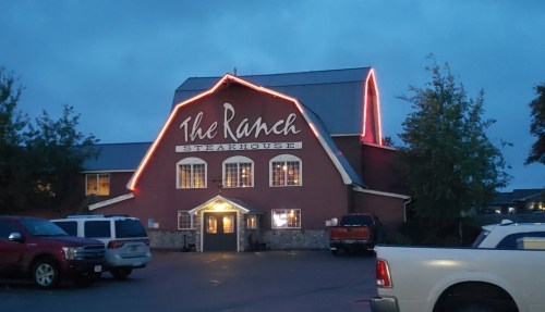 A rustic steakhouse with a red barn-style exterior, illuminated by lights at dusk, surrounded by parked cars.