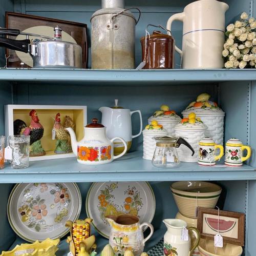 A display of vintage kitchenware and decorative items on blue shelves, including teapots, jars, and framed art.