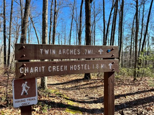 Wooden signpost in a forest directing to Twin Arches (7 miles) and Charit Creek Hostel (1.8 miles) with a clear blue sky.