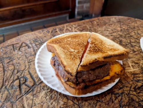 A toasted sandwich with a beef patty and melted cheese, served on a paper plate on a wooden table.
