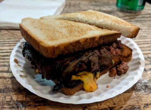 A hearty sandwich with layers of grilled meat and melted cheese, served between two slices of toasted bread on a paper plate.
