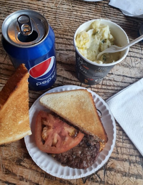 A plate with a sandwich containing a beef patty and tomato, a cup of mashed potatoes, and a can of Pepsi.