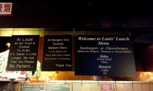 Menu signs at Louis' Lunch, detailing cooking times and burger options, with a welcome message and side choices.