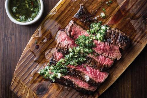 Grilled steak sliced and topped with green sauce, served on a wooden board with a small bowl of sauce on the side.