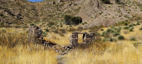 Ruins of a stone structure surrounded by tall grass and rocky hills under a clear blue sky.