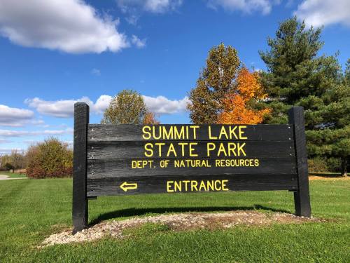 Sign for Summit Lake State Park with "Entrance" arrow, surrounded by trees and a blue sky with clouds.