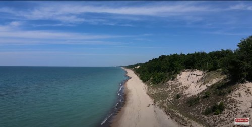 Aerial view of a sandy beach along a calm lake, bordered by lush green trees under a clear blue sky.