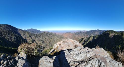 Panoramic view of mountains and valleys under a clear blue sky, showcasing autumn foliage in the distance.