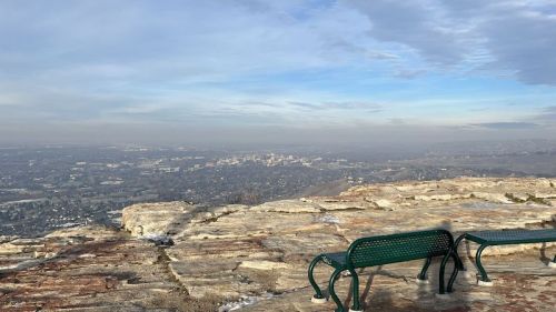 A scenic view from a rocky overlook, featuring two green benches and a hazy cityscape in the distance.
