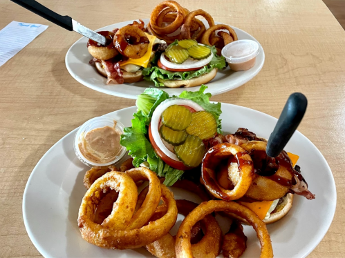 Two plates of burgers topped with bacon, cheese, pickles, and onion rings, served with dipping sauce on the side.