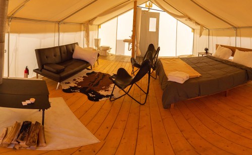 Cozy glamping tent interior with a bed, sofa, chairs, and wooden floor, featuring a rustic decor and a small stove.