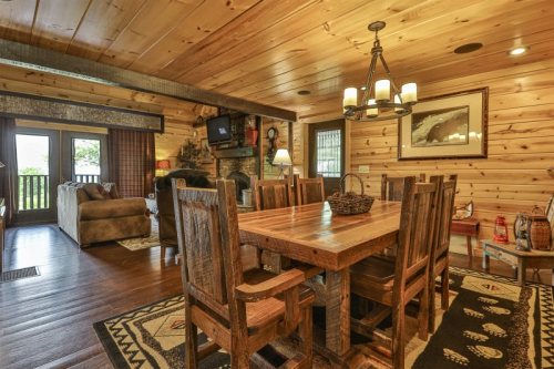 Cozy wooden dining area with a large table, chairs, and a living space featuring a couch and a stone fireplace.
