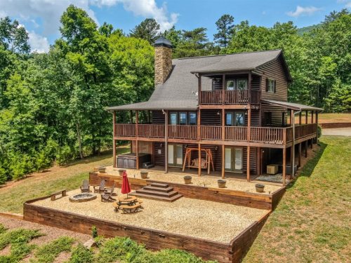 A large wooden cabin surrounded by trees, featuring a spacious deck, outdoor seating, and a fire pit area.