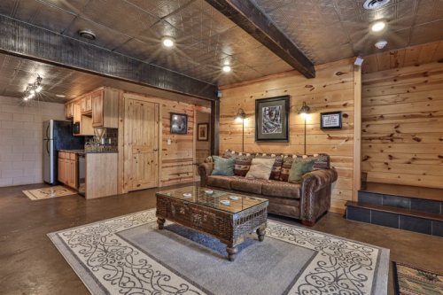 Cozy living room with wooden walls, a leather sofa, coffee table, and a kitchenette in the background. Warm, inviting atmosphere.