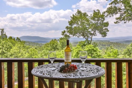 A scenic view of mountains and trees with a table set for wine, featuring a bottle and glasses surrounded by grapes.