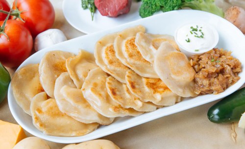 A platter of golden-brown dumplings served with a side of sour cream and caramelized onions, surrounded by fresh vegetables.