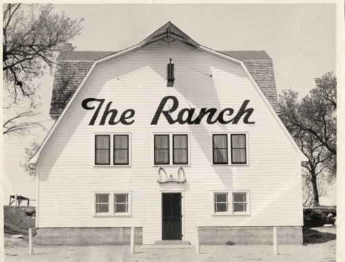 A large white barn with "The Ranch" painted on the front, featuring multiple windows and a door.