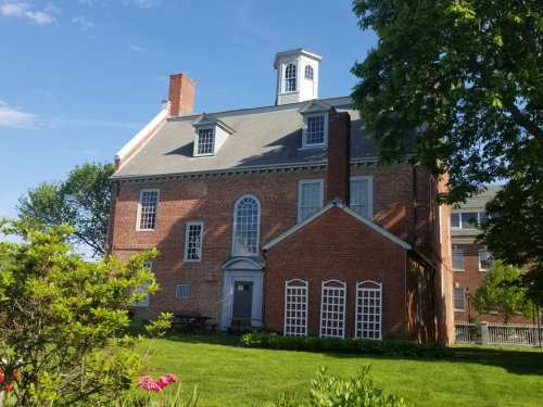 Historic brick building with a gabled roof, large windows, and a cupola, surrounded by green grass and trees.