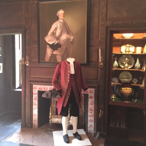 A historical room featuring a portrait and a mannequin dressed in 18th-century attire near a fireplace and display cabinet.