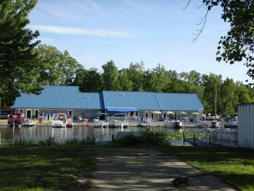 A marina with a blue-roofed building and several boats docked along the water, surrounded by trees.