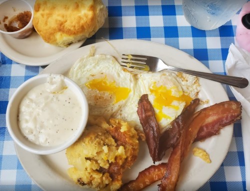 A plate of breakfast featuring two sunny-side-up eggs, bacon, biscuit, and a side of creamy gravy.