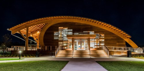 A modern wooden building with a curved roof, illuminated at night, surrounded by green grass and pathways.