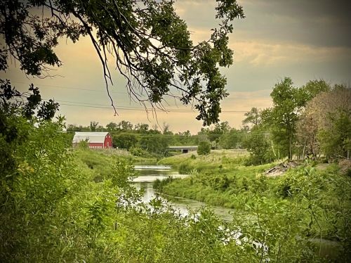 A serene landscape featuring a winding river, lush greenery, and a red barn in the distance under a cloudy sky.