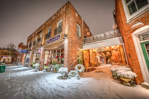 Charming brick buildings in a snowy alley, adorned with festive decorations and warm lights, creating a cozy winter scene.